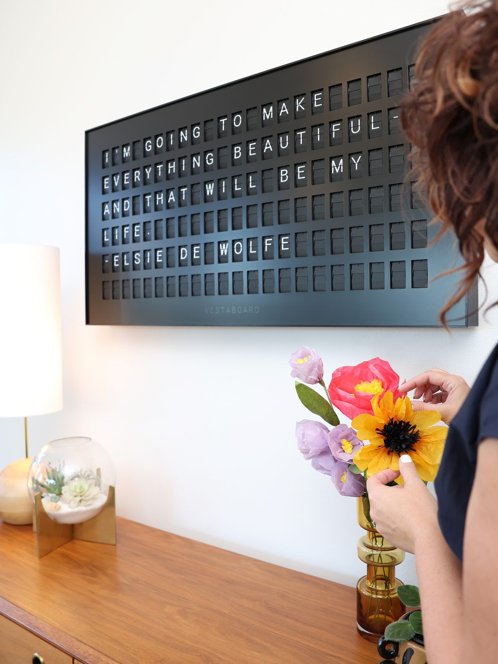 A Vestaboard reading &ldquo;I&rsquo;m going to make everything beautiful and that will be my life. — Elsie de Wolfe&rdquo;, with Sarah arranging paper flowers on the credenza below