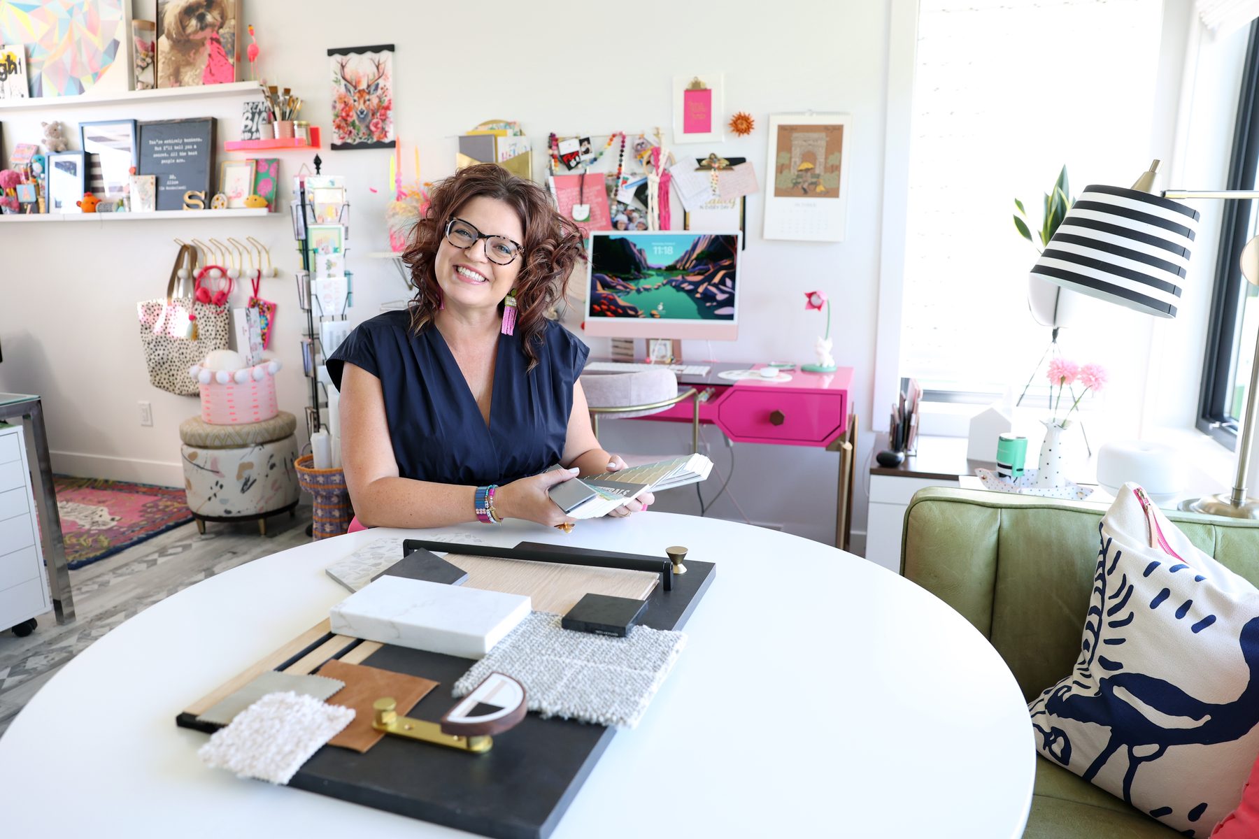 Sarah at the round white table in her studio, a material board in front of her, pink desk and green sofa behind